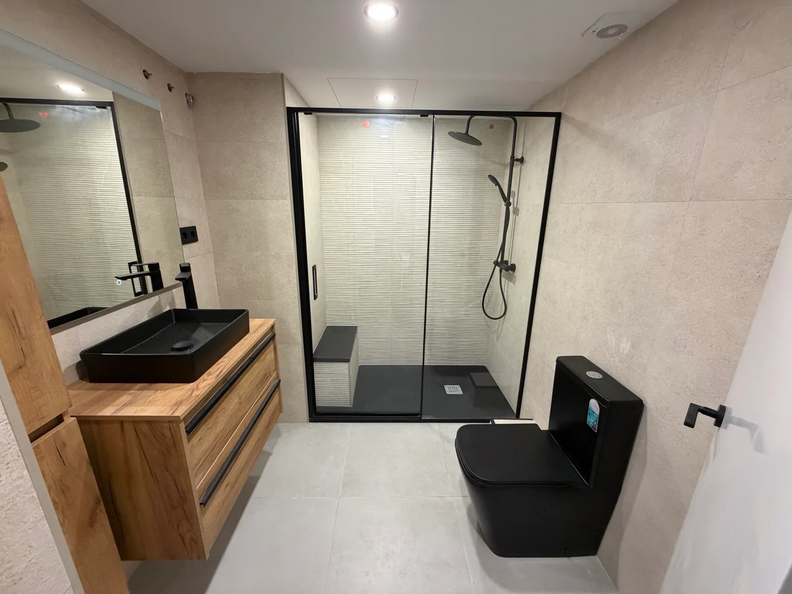 Baño con ducha acristalada de perfilería negra, lavabo negro y mueble de madera.