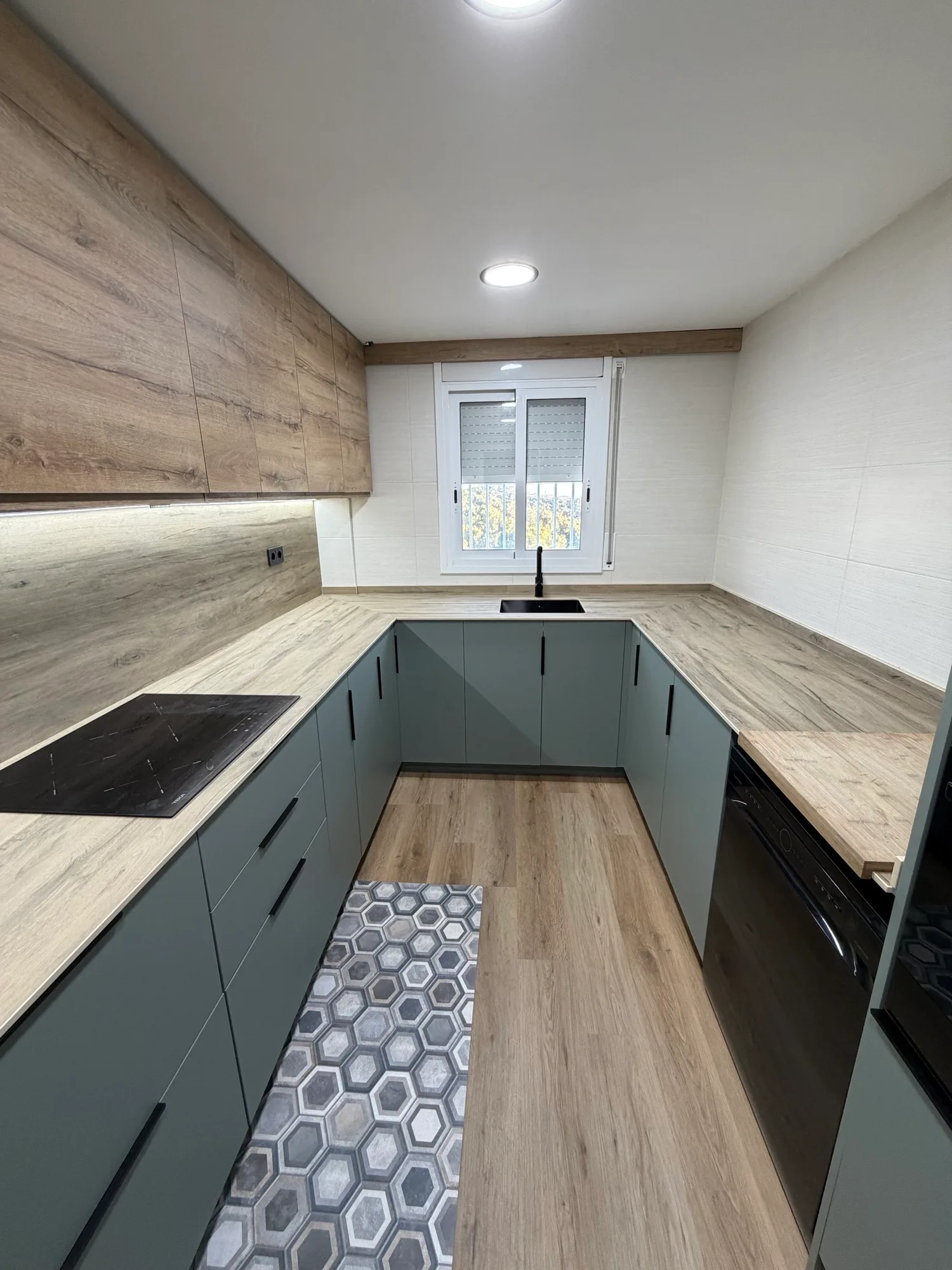 Cocina en U con muebles grises, encimera de aspecto madera, ventana y suelo de dibujo geométrico.